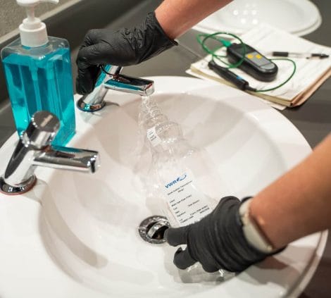 Gloved technician filling a labelled VWR water sample bottle at a basin tap for Legionella microbiological analysis